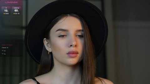 Media: Video of a young woman with fair skin, straight brown hair, and a black wide-brimmed hat, wearing a black top, standing indoors against a blurred background.