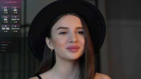 Media: Video of a young woman with fair skin, straight brown hair, and a black hat, wearing a black top, against a dimly lit, modern indoor background.