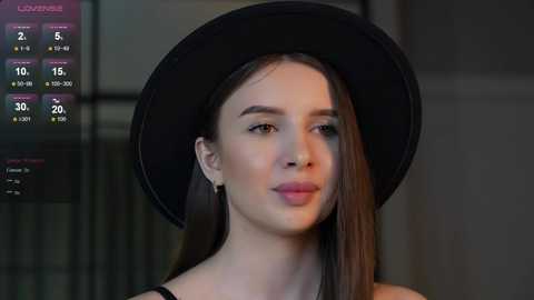 Media: Video of a young woman with fair skin, straight brown hair, wearing a black hat and black top, standing against a dimly lit, blurred background.