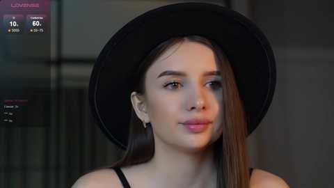 Media: Video of a young woman with fair skin and long brown hair, wearing a black hat and black top, in a dimly lit room with a blurred background.