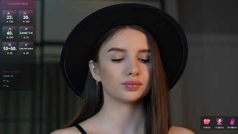 Media: Video of a young Caucasian woman with fair skin, straight brown hair, and closed eyes, wearing a black hat and black top, indoors.