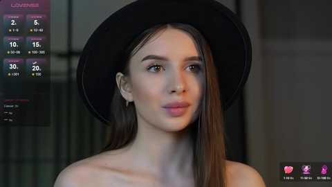 Media: A video of a fair-skinned woman with long brown hair, wearing a black hat and a strapless black top, standing indoors.