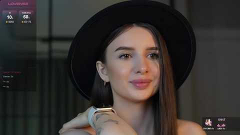 Media: Video of a young woman with long brown hair, wearing a black hat and a light-colored top, smiling softly. She has a tattoo on her right wrist. Background shows a modern indoor setting with blurred windows.