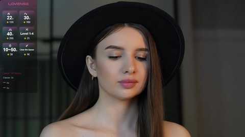 Media: Video of a young woman with fair skin and straight brown hair, wearing a black hat and a black top, against a blurred indoor background.