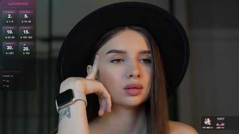 Media: Video of a young woman with fair skin, long straight brown hair, and a black hat, wearing a black top, looking contemplative with a hand on her cheek, in a dimly lit room.