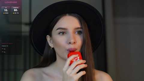 Media: Video of a young woman with long brown hair, wearing a black hat and drinking from a red bottle, set against a dark background.