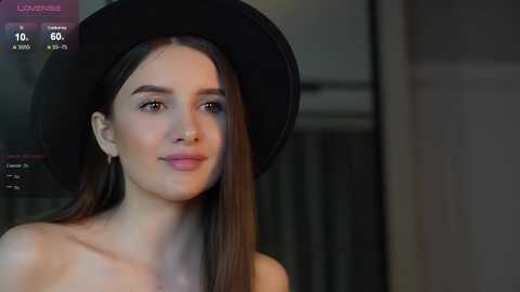 Media: Video of a young woman with long brown hair, wearing a black hat and light makeup, standing against a blurred background.