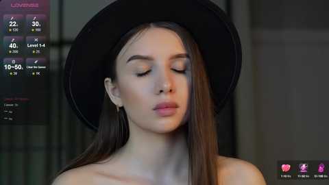 Media: Video of a young woman with long brown hair, wearing a black hat, closed eyes, and subtle makeup, set against a dimly lit, modern backdrop.