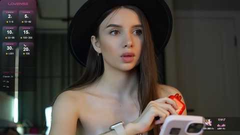 Media: A video of a young woman with long brown hair, wearing a black hat and a red lipstick, holding a phone, taken indoors.