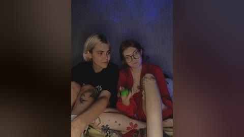 Video of two women sitting on a bed, one blonde with a black shirt, the other with glasses and a red top, in a dimly lit room.