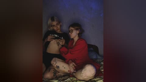 Video of two women in a dimly lit room; one with a tattooed belly and short blonde hair, wearing a black crop top, the other with glasses and a red sweater.