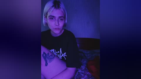Video of a young, androgynous person with short platinum blonde hair, wearing a black graphic t-shirt with a stylized logo, sitting on a bed with patterned sheets. The background features a dimly lit room with a purple hue.