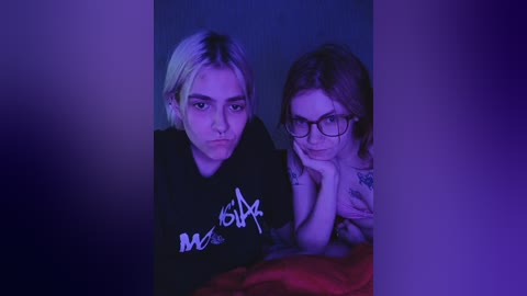 Video of two women with pale skin, one with short blonde hair and glasses, the other with brown hair, leaning on each other, bathed in purple-blue light, wearing black clothes.