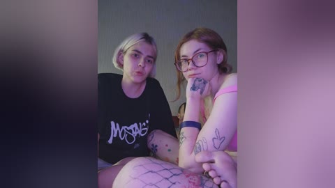 A video featuring two young women with tattoos, one in a black t-shirt and the other in glasses and pink lingerie, sitting close together against a gray wall.