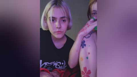 Video of a young woman with short blonde hair, wearing a black t-shirt, and a man with tattoos, both with serious expressions.