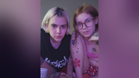 Video of two women with tattoos, one blonde with a black t-shirt and the other with glasses, sitting close together.