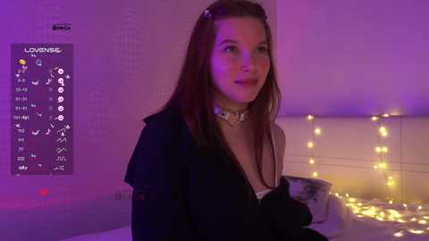 Video of a young woman with long red hair, wearing a black jacket, sitting on a bed with white bedding and yellow string lights. Purple ambient lighting.