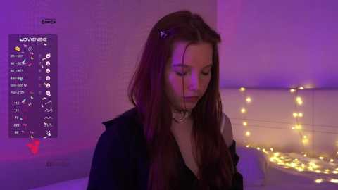 Video of a young woman with long brown hair, wearing a black jacket, standing in a dimly lit room with purple lighting, a string of fairy lights, and a white wall.