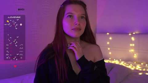 Video of a young woman with long, straight brown hair, wearing a black off-shoulder top, standing in a bedroom with purple lighting and a calendar poster, holding her collar.