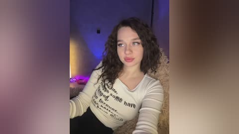 Video of a young woman with curly dark hair, wearing a white crop top with text, sitting on a fur rug in dimly lit room, with candles and purple lighting.