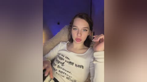 Video of a young woman with fair skin, wavy brown hair, and full lips, wearing a white top with black text. She is seated on a beige chair, indoors with dim lighting.
