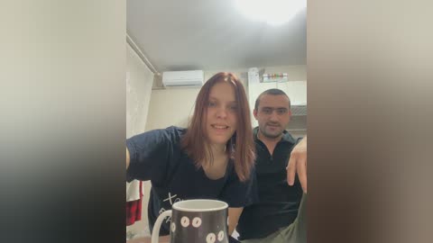 A video of a smiling woman with shoulder-length red hair, wearing a navy blue t-shirt, and a man with short dark hair in a dark shirt, standing in a small, dimly lit room with white walls and an air conditioner.