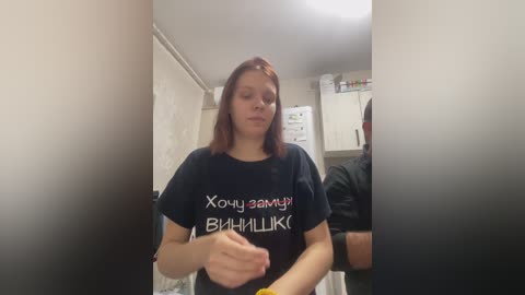 Video of a young Caucasian woman with shoulder-length red hair, wearing a black T-shirt with Cyrillic text, standing in a dimly lit kitchen.