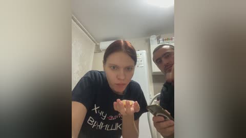 A video of a woman with auburn hair in a navy T-shirt, holding a slice of pizza in her mouth, standing beside a man with a beard in a dimly lit kitchen.
