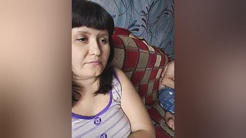 A video of a young woman with short black hair and a white and purple striped top lying on a bed with a red and beige patterned pillow, next to a baby wearing a blue and white striped outfit.