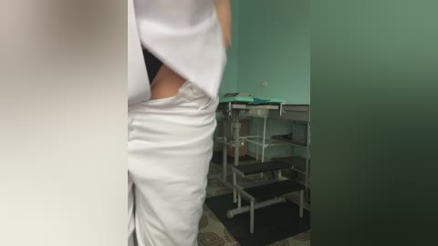 Media: Video of a person in white scrubs and a mask, partially obscured by a blurred object, standing in a medical room with green walls and metal examination tables.