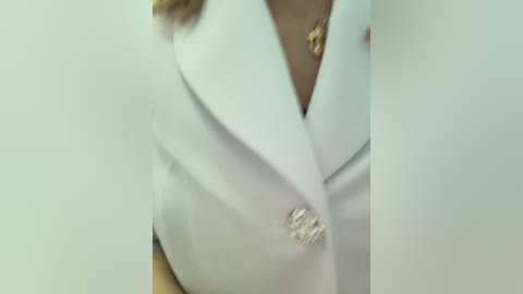 Media: A video showing a close-up of a woman's upper torso, wearing a white blazer with a gold brooch on the left lapel, and a gold necklace. The background is a plain, light-colored wall.