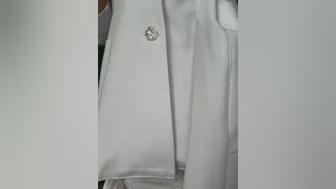 Media: A close-up video of a white lab coat with a small, circular, silver emblem on the left lapel, indicating a medical or scientific professional's attire. The coat is neatly folded, revealing its smooth, crisp texture.