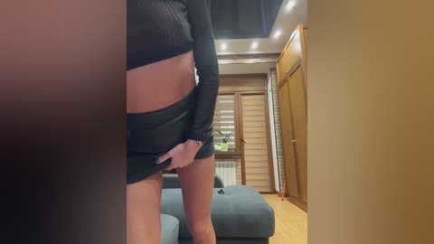 Video of a slender woman with light skin, wearing a black mesh crop top and shorts, standing in a modern, beige-toned room with wooden furniture and a blue couch.