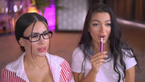 Video of two young women, one with glasses, the other with long dark hair, eating lollipops in a modern room with purple lighting.