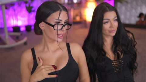 Video of two women with long black hair, one in glasses, wearing black tops, indoors with purple lighting, blurred background.