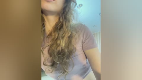 Video of a woman with light skin, long wavy brown hair, wearing a pink T-shirt, partially visible face, blurred background with white walls.