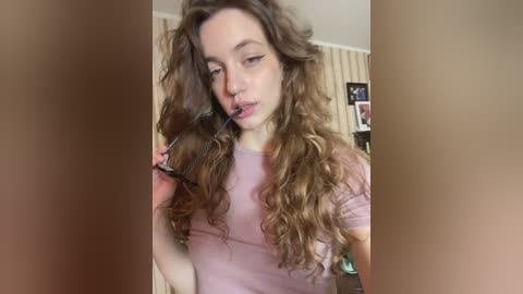 Video of a young woman with long, curly brown hair, wearing a pink shirt, playfully holding glasses in front of her face, indoors with beige wallpaper and framed pictures in the background.