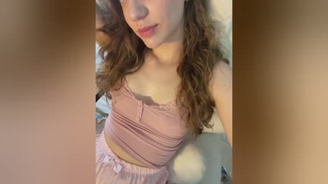 Video of a young Caucasian woman with light skin and long, wavy brown hair, wearing a light pink, lacy camisole and matching pants, sitting on a white chair.