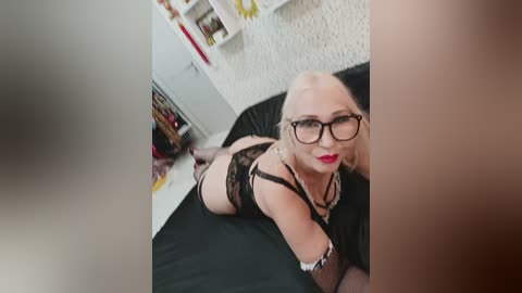 A video of a fair-skinned, blonde woman with glasses, wearing black lingerie and thigh-high stockings, lying on a black bed in a bedroom with white walls and shelves.