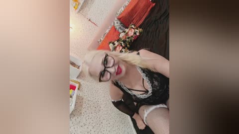 Video of a blonde woman with glasses in a black lace maid outfit, sitting on a bed with floral pillows, in a speckled room.