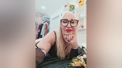 A video of a blonde woman with glasses and red lipstick, wearing a black top, lying on a bed, with a closet and shelves in the background.