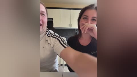 Media: A video of two people in a kitchen. A man with short hair and a white jacket with black stripes is laughing. A woman with long dark hair and a black shirt is hiding her mouth with her hand.