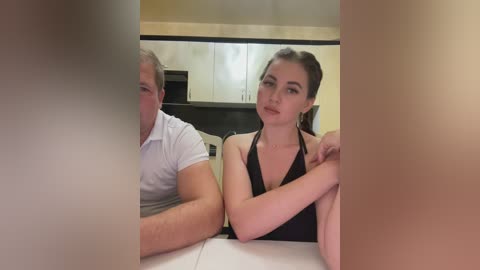 Media: Video of a young woman with fair skin and brown hair tied back, wearing a black halter top, sitting next to an older man with gray hair and a white shirt, in a modern kitchen with light-colored cabinets and a black countertop.