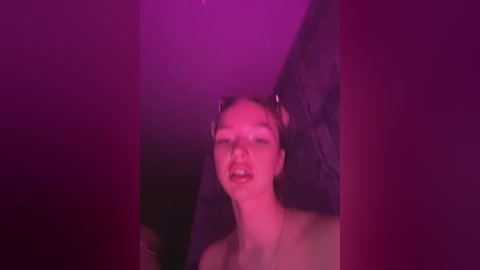 Media: Video of a young, fair-skinned woman with dark hair, wearing a black lace dress, standing against a dark background, bathed in vibrant pink and purple lighting, creating a surreal, dreamlike atmosphere.