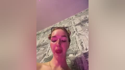 Media: Video of a fair-skinned woman with light brown hair styled in a bun, wearing dark sunglasses and pink lipstick, making a pouty face against a backdrop of a grayscale New York City skyline mural.