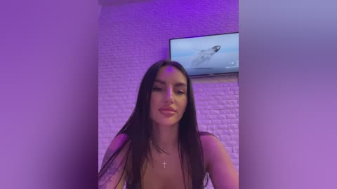 Media: Video of a young woman with long dark hair, wearing a white top, standing in a room with a brick wall and a TV showing a rocket launch, under purple lighting.