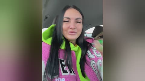 Video of a young woman with long black hair, wearing a pink hoodie with neon green accents, smiling, sitting in a car with blurred background.