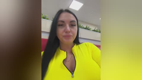 Video of a woman with long black hair, fair skin, and a medium build, wearing a bright yellow zip-up hoodie, smiling slightly. Background features a modern indoor setting with a white ceiling and green plants.