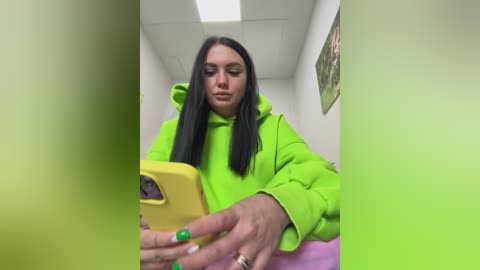 Video of a young woman with long black hair, wearing a bright green hoodie, looking at a yellow smartphone. The background features a white wall with a framed picture and fluorescent lighting.