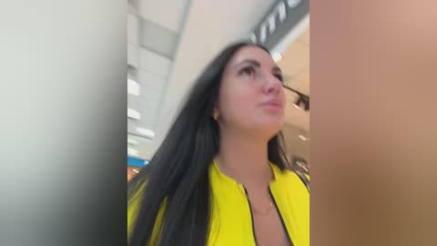 Video of a woman with long black hair in a yellow jacket, standing in a brightly lit indoor setting.